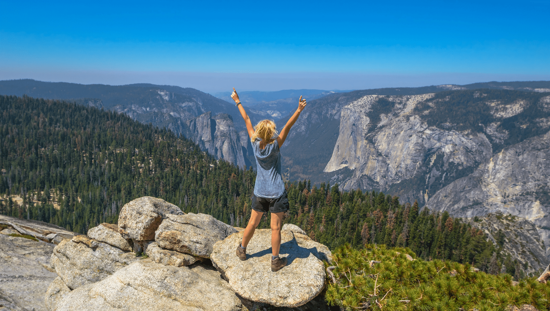 A female hiker stands on a large rock in a valley, surrounded by mountains in Yosemite National Park. She has her hands held high above her head in awe of the view.