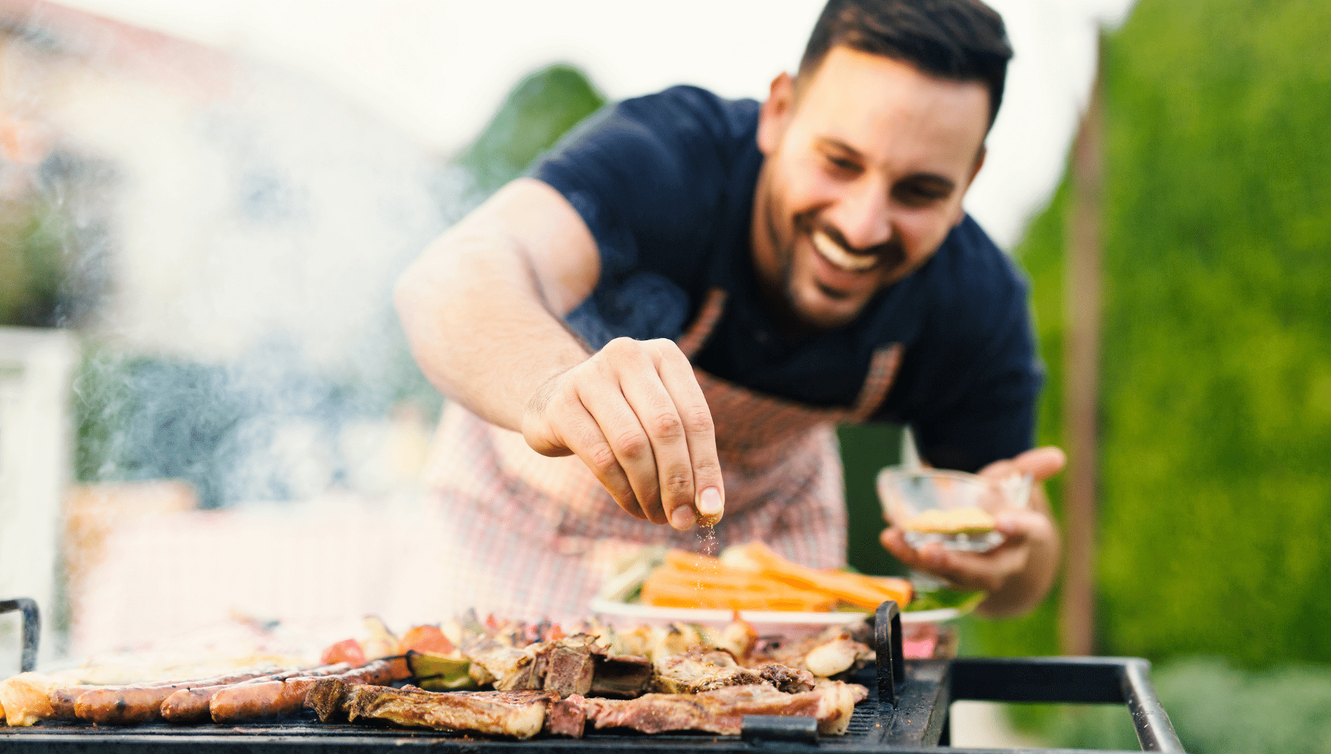 A smiling man leans over the grill as he adds seasoning to his food. Smoke is coming up from the grill.