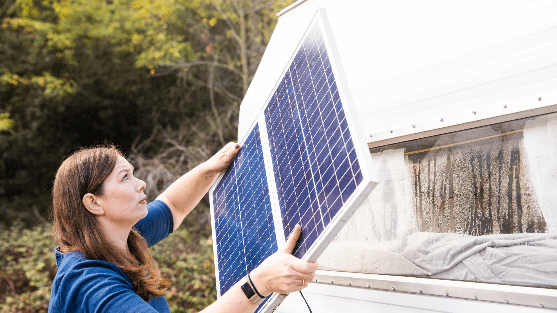 Woman with dark hair and blue shirt looks kinda confused as she attempts to install a solar panel on her RV. Her RV has dirty windows, but I don't think that's her main concern. She is likely installing the solar panel so she can power her RV's appliances and air conditioner.