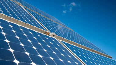 A group of blue solar panels reflect the sun with a blue sky in the background. What is better to connect solar panels in a series or parallel?