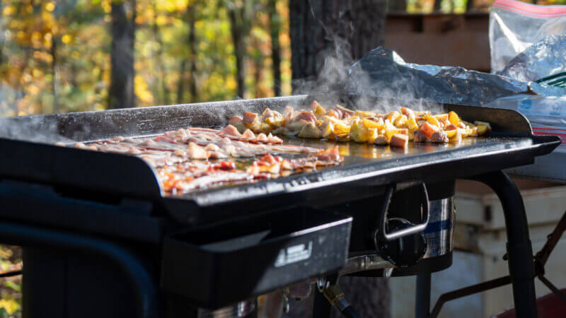 A blackstone griddle is topped with breakfast classics and makes the best meals! This one is being used at an RV campsite outside in the woods.