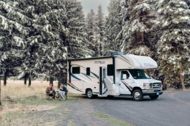 Two people sit outside their class C RV in early winter to enjoy the snow covered trees.