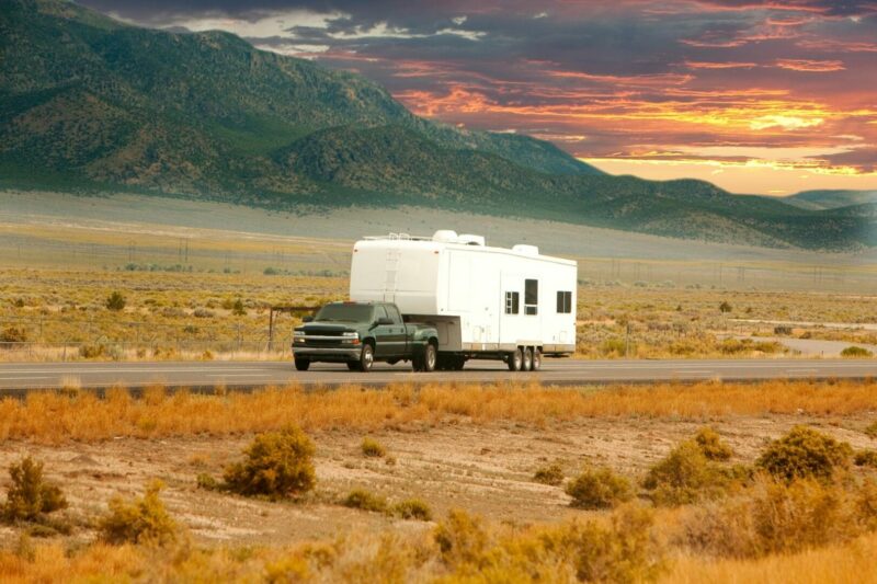 Truck towing a 5th wheel trailer down a road with mountains and a sunset in the background.