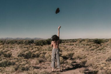 A woman with her back to the camera throws off her top in the desert.