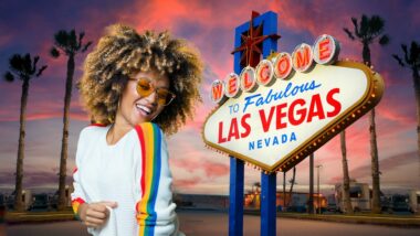 An ethusiastic woman with colored glasses and afro grooves next to a sign welcoming you to Las Vegas in front of an RV campground at sunset