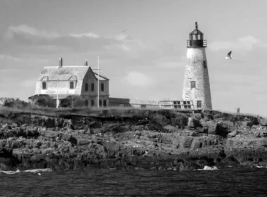 A black and white photo taken of the the abandoned Wood Island Lighthouse in Maine with growth on the tower and dirty shingles covering the attached house.