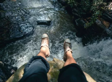 A woman hangs her feet over the edge of a waterfall as she sits on a boulder.