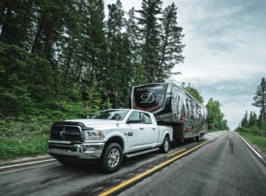 White truck pulling DRV Toy Hauler on the road with forest in the background.