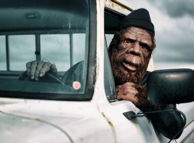 Person dressed up as bigfoot driving a white pickup truck as they lean out the window.