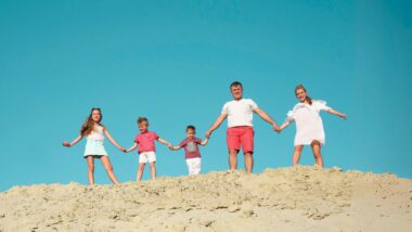 A photo of a family of five holding hands while standing on a sandy hilltop with a bright blue sky in the background.