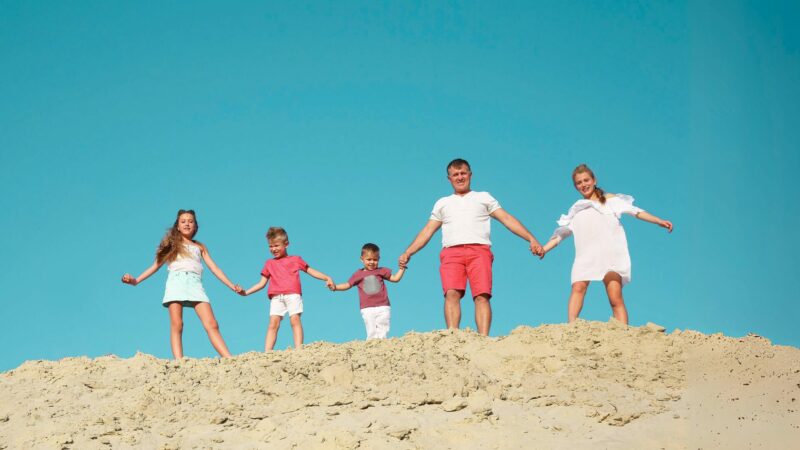 A photo of a family of five holding hands while standing on a sandy hilltop with a bright blue sky in the background.