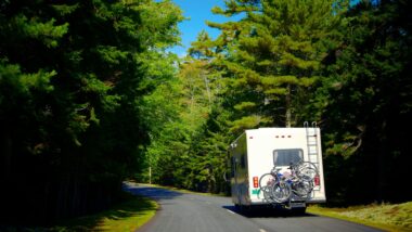 An RV driving down a heavily wooded road with bikes secured to the rear of it.