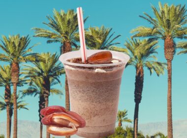 A shake with dates upon and beside it as it sits in front of a photo of date trees side by side with a bright blue background.