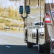 A motorhome towing a vehicle behind it on a road with trees colored with fall leaves in the background.