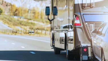 A motorhome towing a vehicle behind it on a road with trees colored with fall leaves in the background.