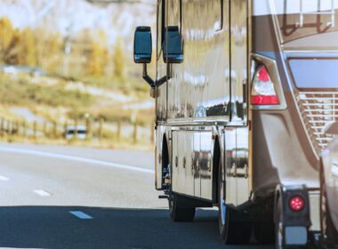 A motorhome towing a vehicle behind it on a road with trees colored with fall leaves in the background.