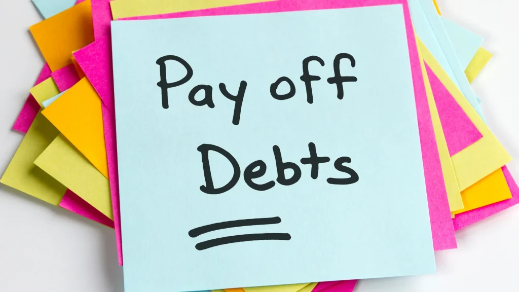 A messy stack of post-it notes in gold, hot pink, yellow, and light blue colors with the top post-it stating "Pay off Debts" with the word "Debts" underlined twice.