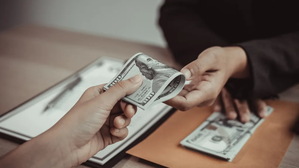 A person exchanging money with another perosn as some documents sit beneath the exchange on a table between the two people.