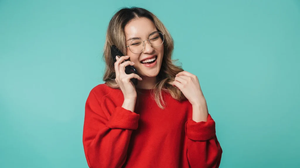 A woman in glasses wearing a red sweater is on the phone as she stands against a teal background.