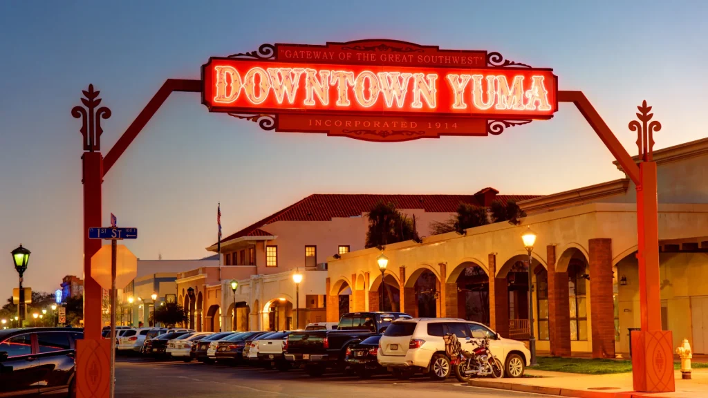 Downtown Yuma and its neon sign, cars are parked down the street along spanish inspired building.