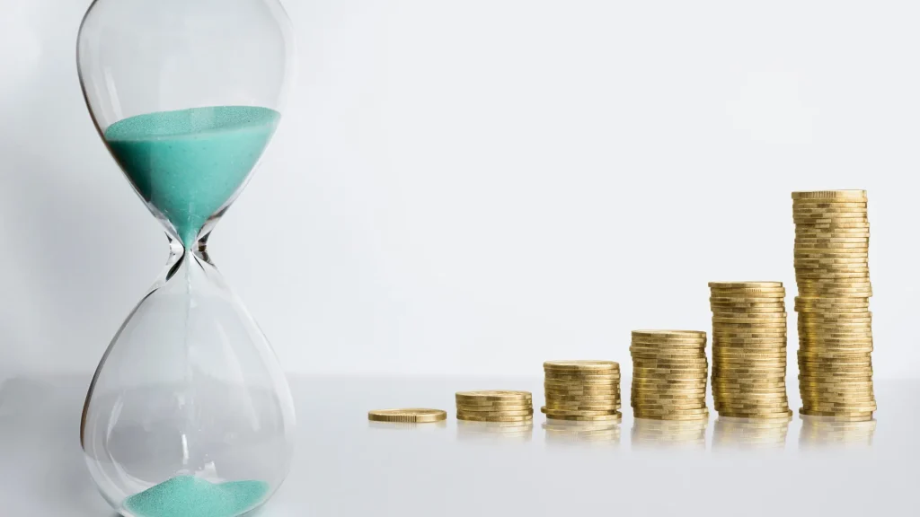 An hourglass with turqoise sand beside a rising stack of change beside it, against a white background.