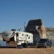 A fifth wheel parked in front of a large rock sitting beside a body of water as a man sits in an outdoor chair beside his RV and his truck sits parked on the opposite side of the RV.