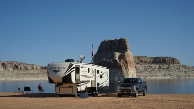 A fifth wheel parked in front of a large rock sitting beside a body of water as a man sits in an outdoor chair beside his RV and his truck sits parked on the opposite side of the RV.