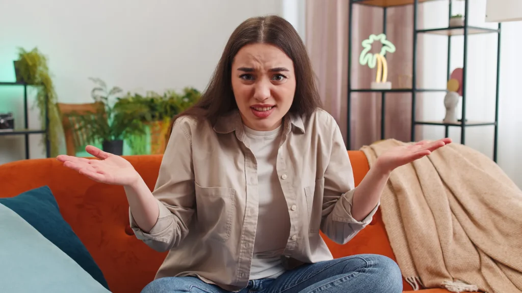 A woman holding her hands up shrugging, gesturing "Why" as she sits atop her burnt orange sofa with decor behind her.