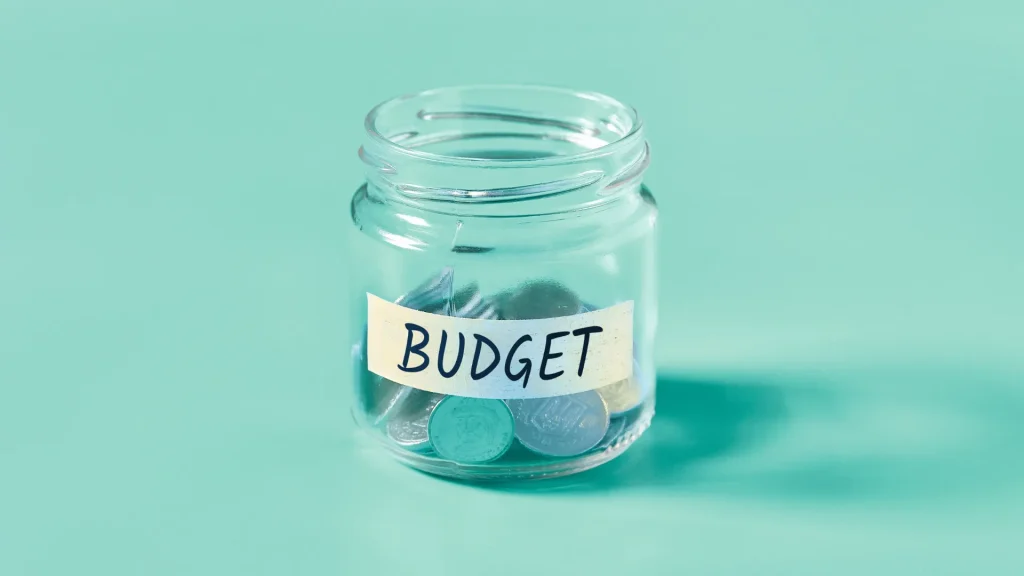 A clear glass jar labled "BUDGET" with coins in it against a teal blue background.