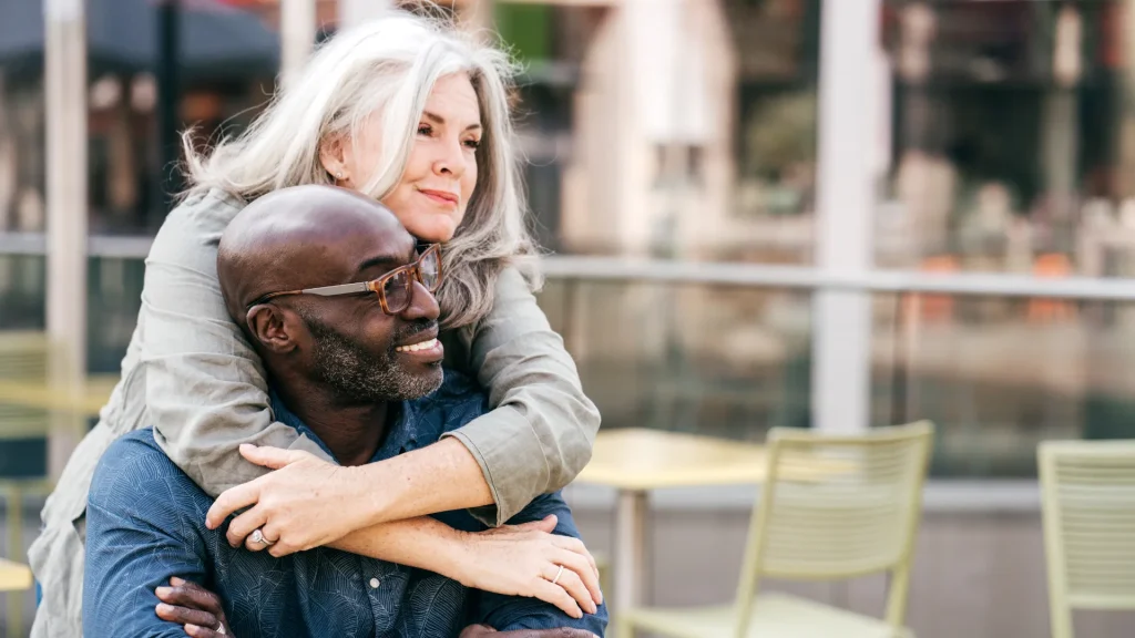 An older couple together in an outdoor seating area as one wraps their arms around the other as they sit.