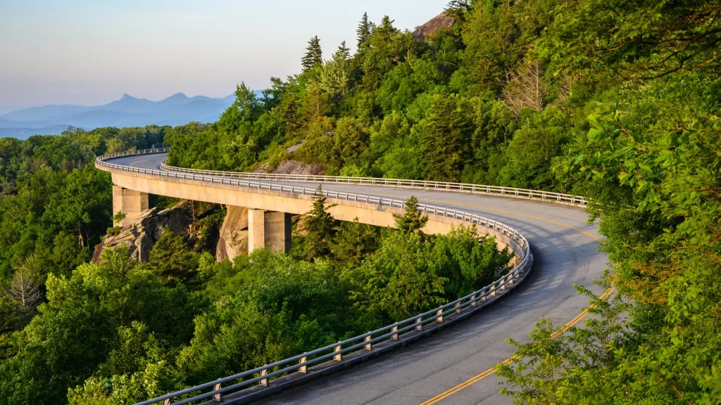 The Blue Ridge Highway in Virginia near Fancy Gap. It's a winding highway around mountains with green foliage hanging off the landscape. More mountains sit in the background far in the distance. 