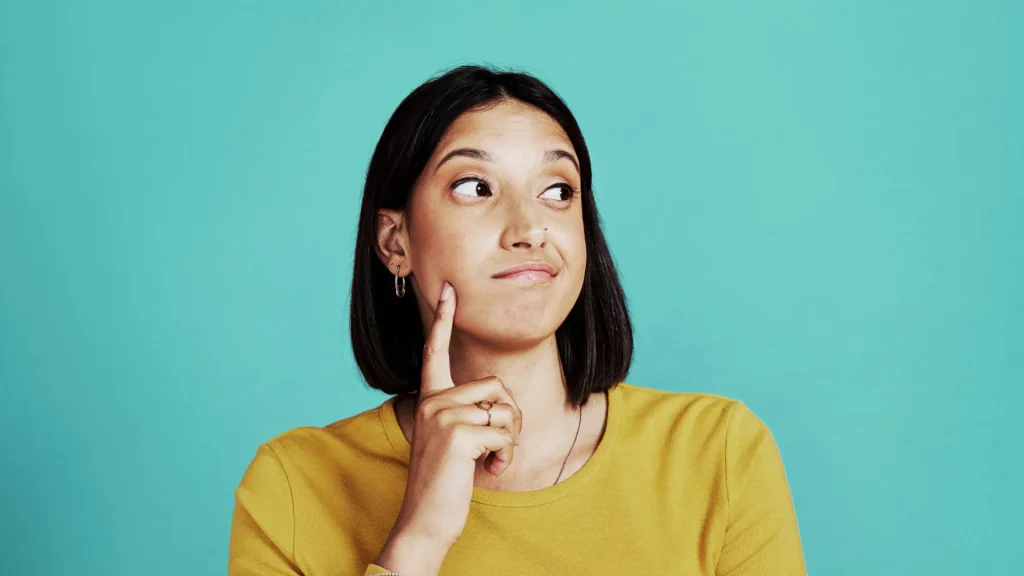 A person in a mustard yellow long sleeve crew shirt holding their finger up to their face as they look off to the side, gesturing that they are thinking about something. This photo is being utilized to emphasize visually someone is thinking about where to put their TV mount.