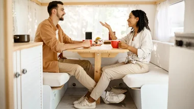 A couple seated in their dinette within their RV as they look at each other and talk, while drinking coffee out of red mugs.