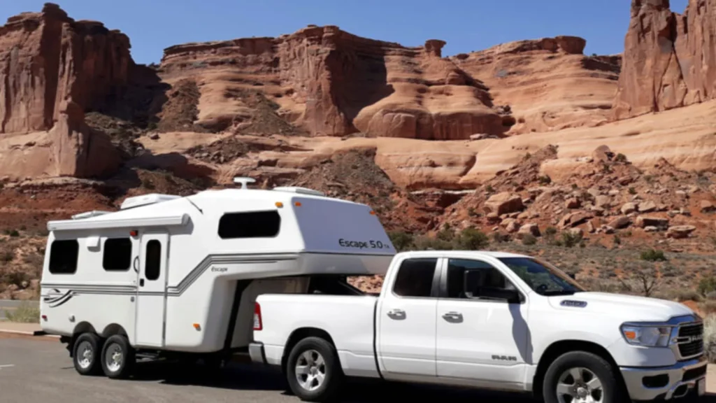A white Dodge Ram pickup truck towing an Escape 5.0 travel trailer. It sits parked along teh side of a road or parking lot with red rocky landscapes behind it. 