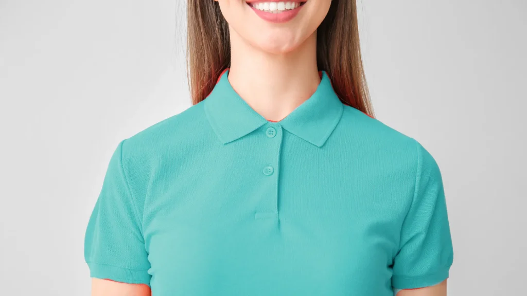 A woman in a teal blue polo smiling against a white wall.