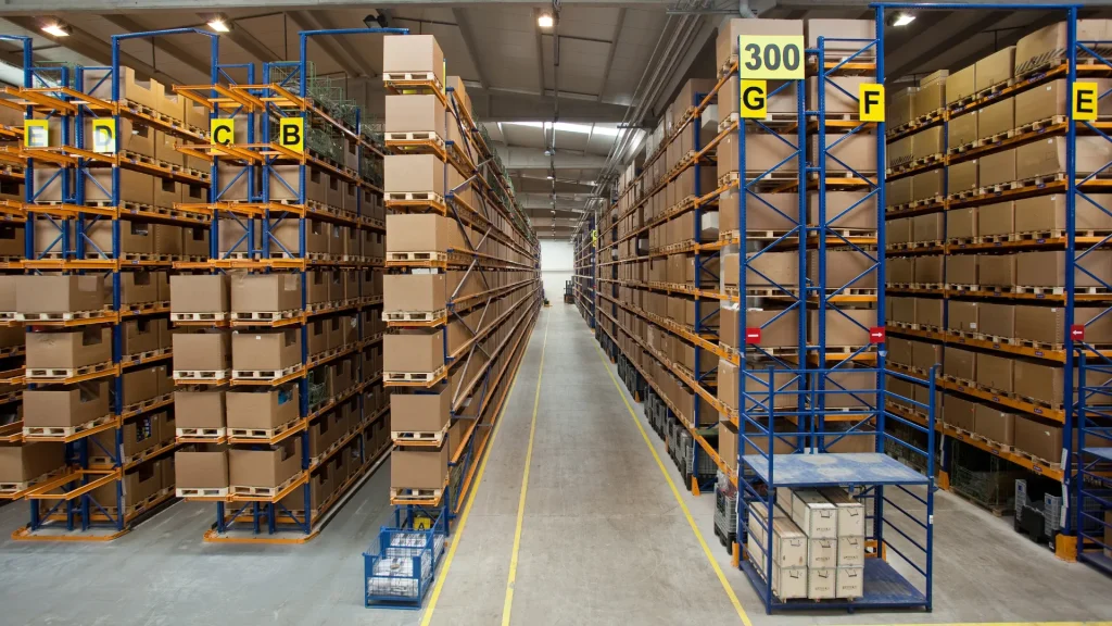 A ware house filled with aisles of boxes with alphabetical yellow labels on each aisle as floor to ceiling blue metal shelving holds up the boxes.