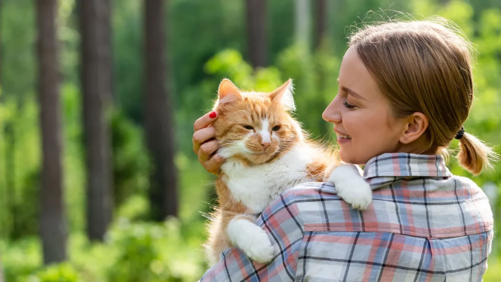 A woman outdoors in a plaid outdoor long sleeve button-up shirt as she holds her long-haird orange and white cat at her shoulder. They're surrounded by green foliage.
