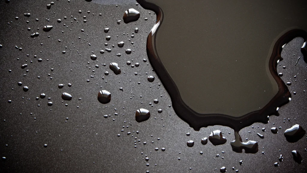 A dark black water and stain resistant surface covered in a liquid that pools in droplets and a puddle atop it.