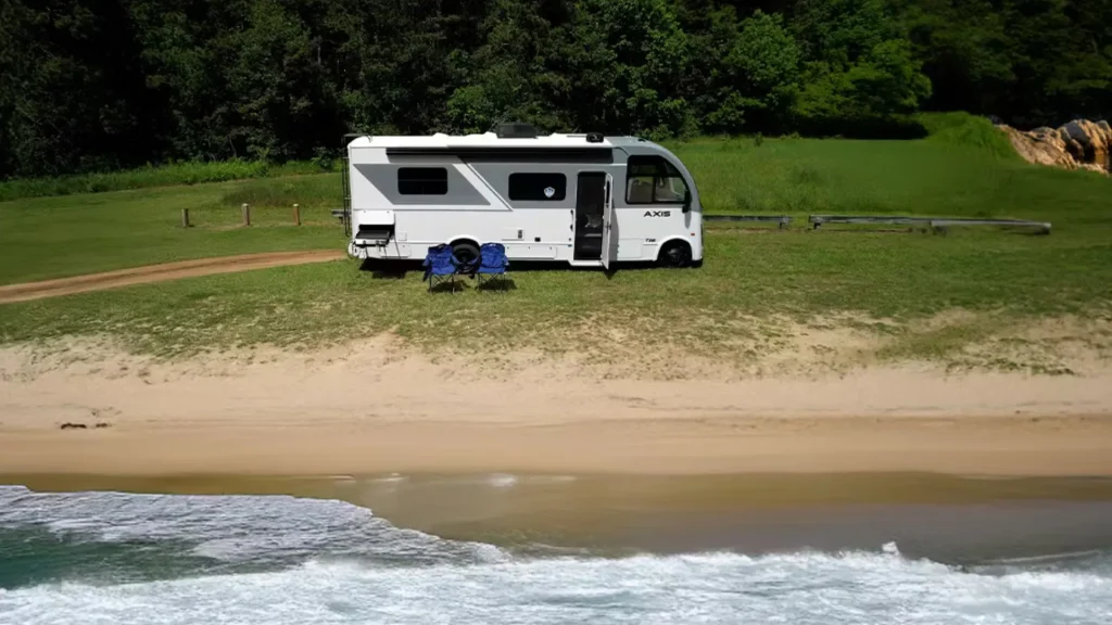 The grey and black exterior of the Thor Axis 24.1 as it sits on a beach with a wood fence behind it along with a dense forest. Two blue camping chairs sit in front of the parked motorhome. 