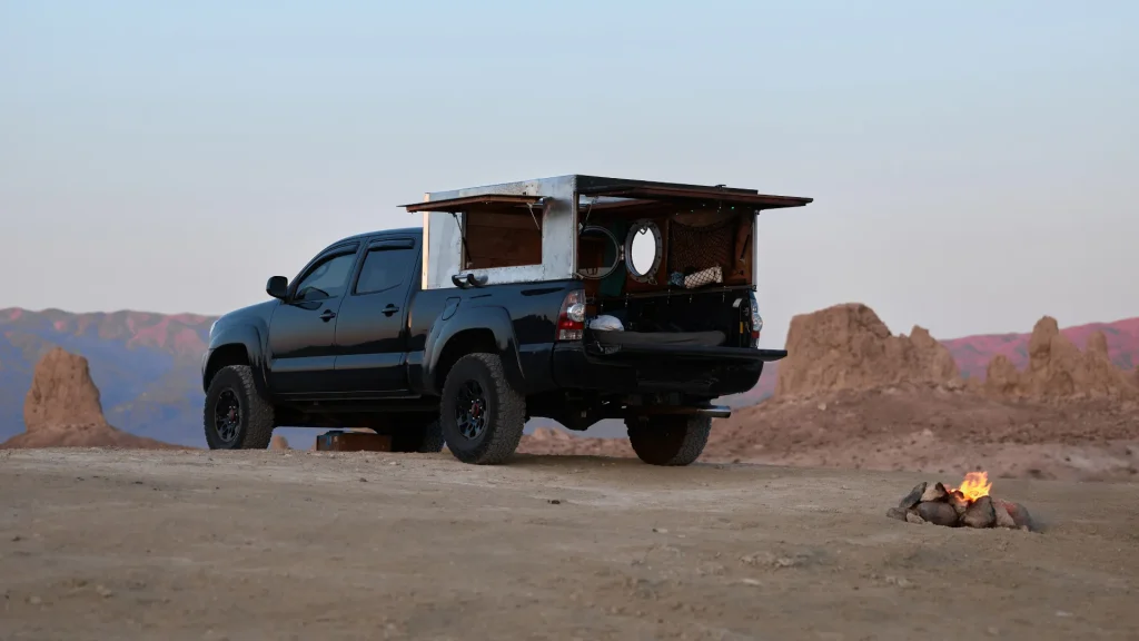 A black pickup truck with a camper in the bed of the truck is parked at the edge of a rocky landscape as the sun sets, painting the rocks and sky in pinks and reds. A small fire is lit besdie the truck. 