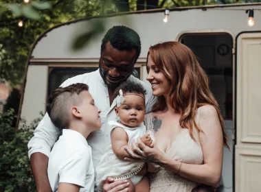 A family of four standing outside their travel trailer for a professional photo as the mother holds a baby, the father looks down at his son.