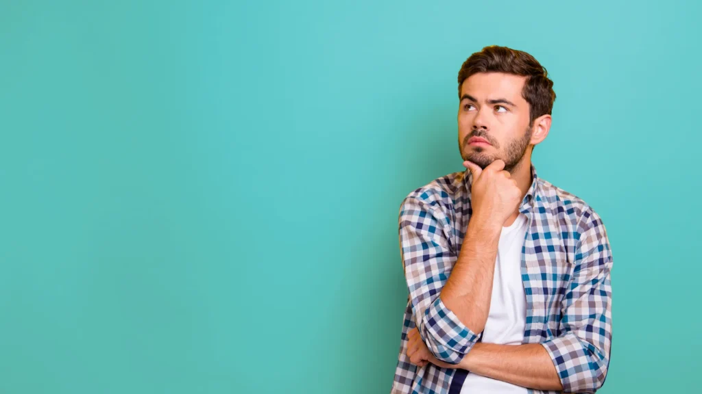 A man holding their hand to their chin as they look upwards quizically as if to be pondering against the teal blue background. He wears a blue plaid dress shirt open with a white t-shirt beneath.