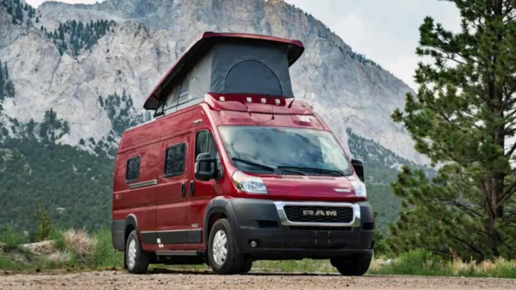 The Winnebago Solis in red, parked in a gravel spot bseides a fir tree as a mountain stands behind it. 