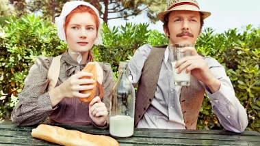 Two people seated side-by-side at a wooden picninc table drinking milk and eating a baguette of bread, representing the Amish Lifestyle.