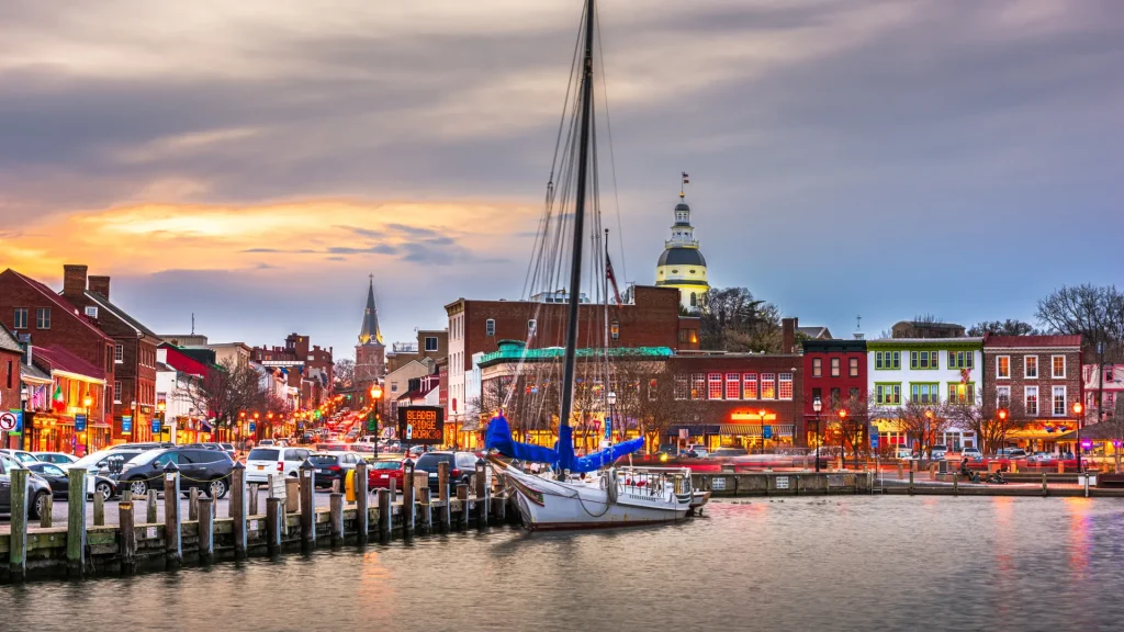 Annapolis Harbor at Dusk with a single sailboat docked. There's a parking lot filled with cars to one side as the city lights give the photo a warm and lively look.