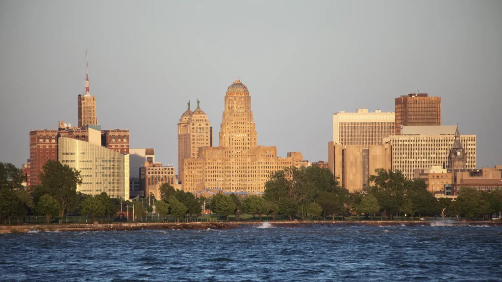 An aerial of Buffalo, NY city skyline witht he water at the foreground of the photo. The water is dark and unsaturated. The buildings are sepia toned and the sky is grey.