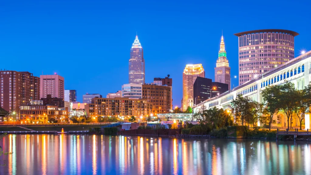 Cleveland, OH at night with the city lights reflecting into the waters. The sky is a deep royal blue and the lights range from ambers, blue whites, and greens.