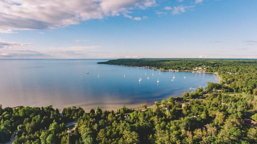 An aerial photo of Ephraim in Door County, WI. The bay is a pretty blue, reflecting the blue sky dotted with clouds. Lush green forests surround the bay as sail boats float effortlessly atop the glass-like surface.