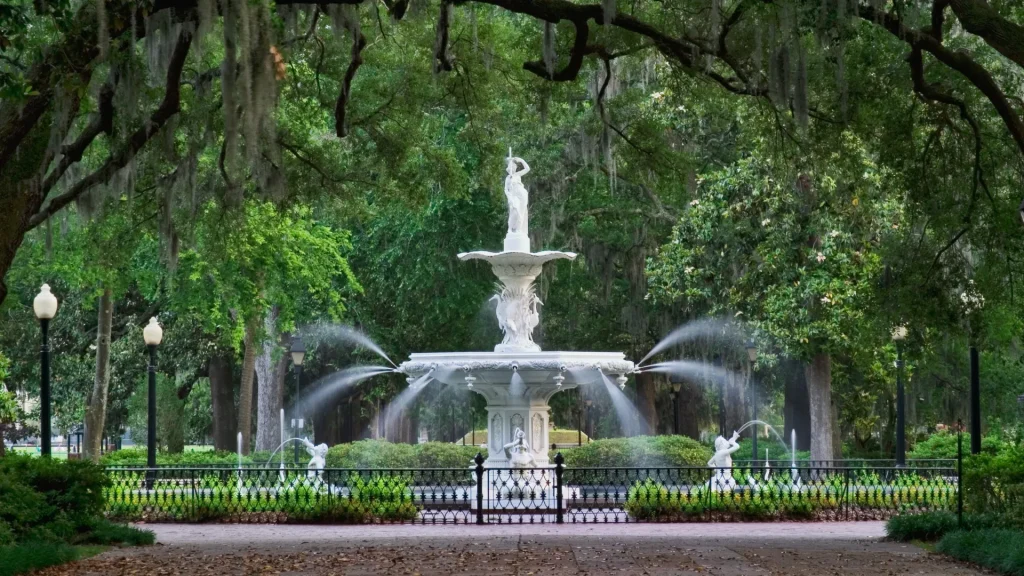 The white Forsyth Park Fountain in Savannah, Georgia surrounded by mature trees, manicured shrubs, and lawns.