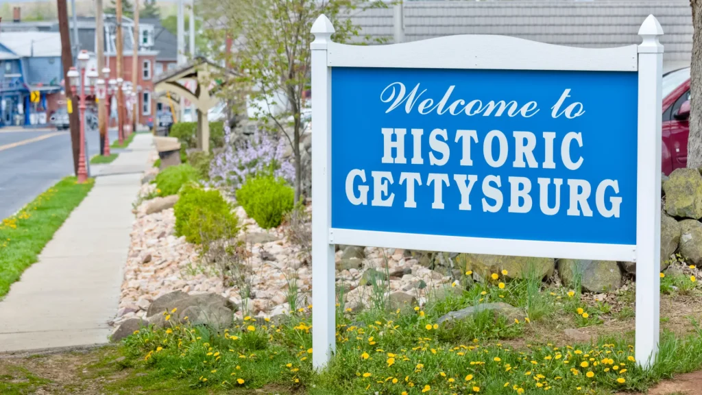 A white frame holds a Carolina blue sign saying "Welcome to Historic Gettysburg". It sits in the grass with dandelions blooming around it. A street and sidewalk pull us into the background of the photo where buildings stand.